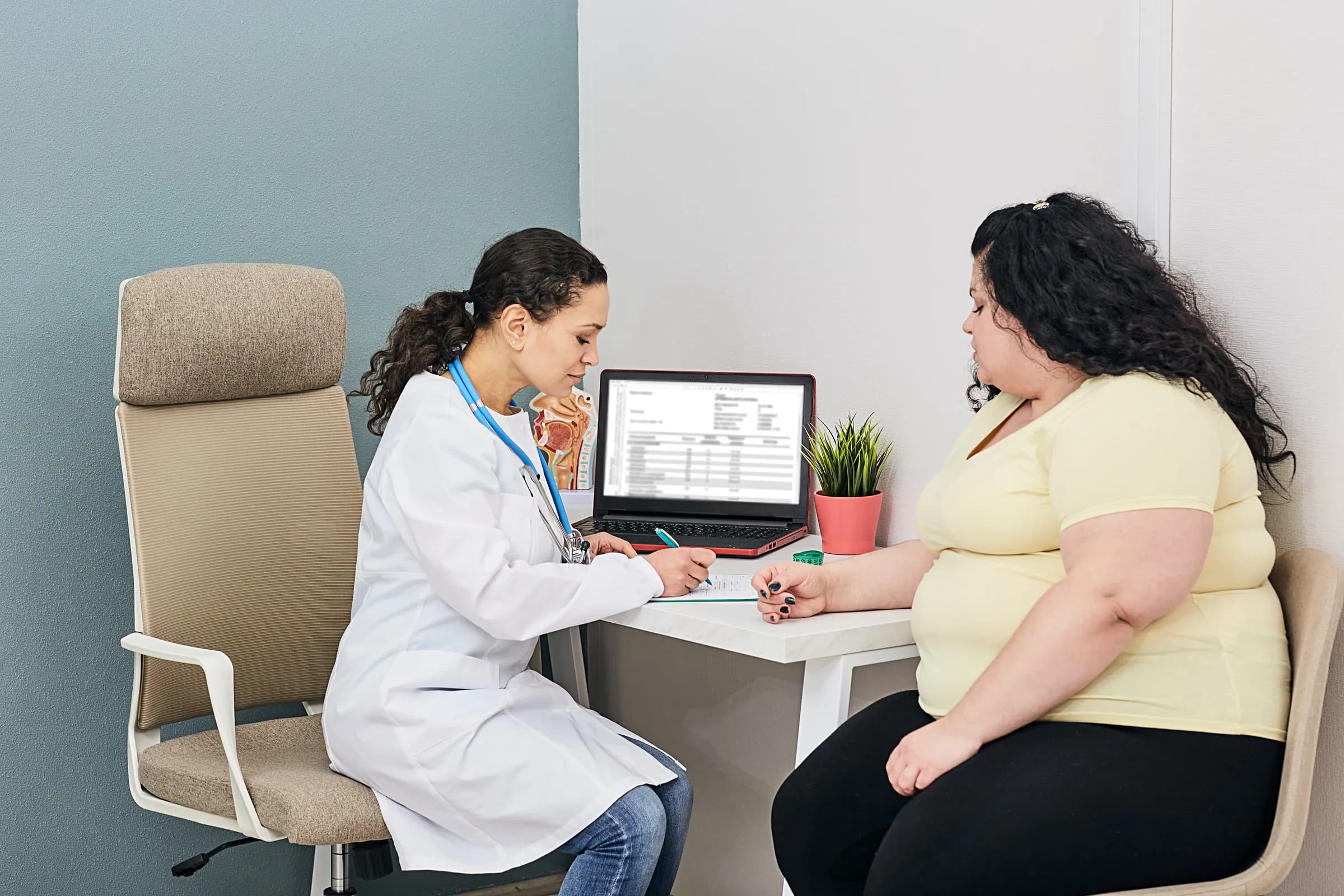 A clinician in a white coat speaks with a woman across a desk in a consultation room