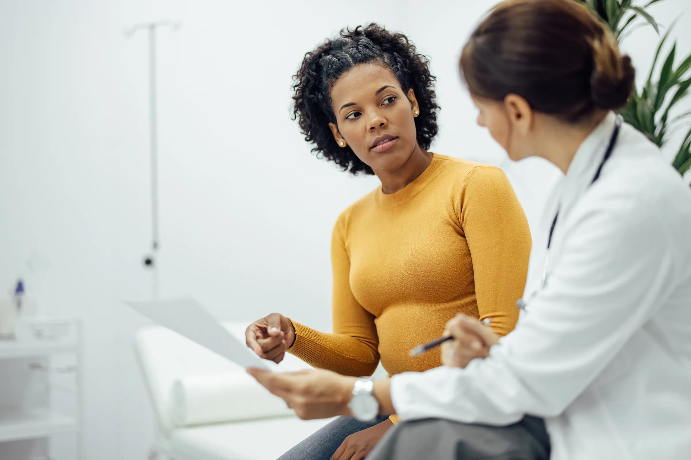 A woman in a mustard sweater holds a sheet of paper while speaking with a clinician in a white coat
